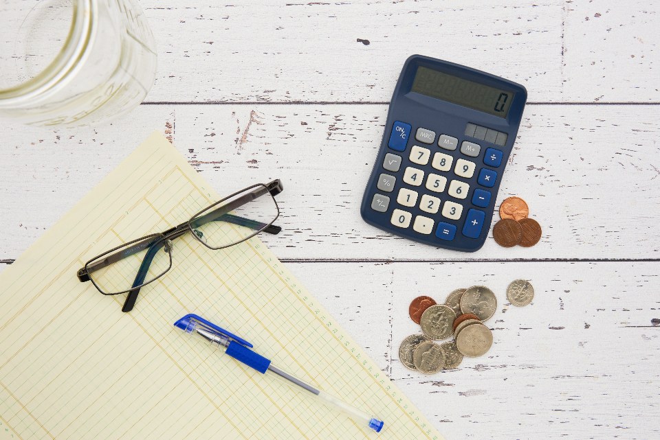 Calculadora, óculos, caneta e moedas sobre papel pautado